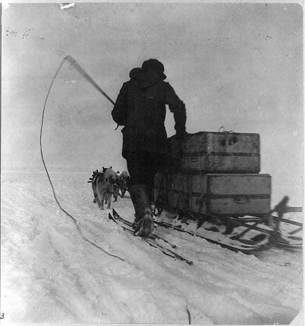 Archival photograph: a man in furs with a whip beside a dog sledge loaded with crates
